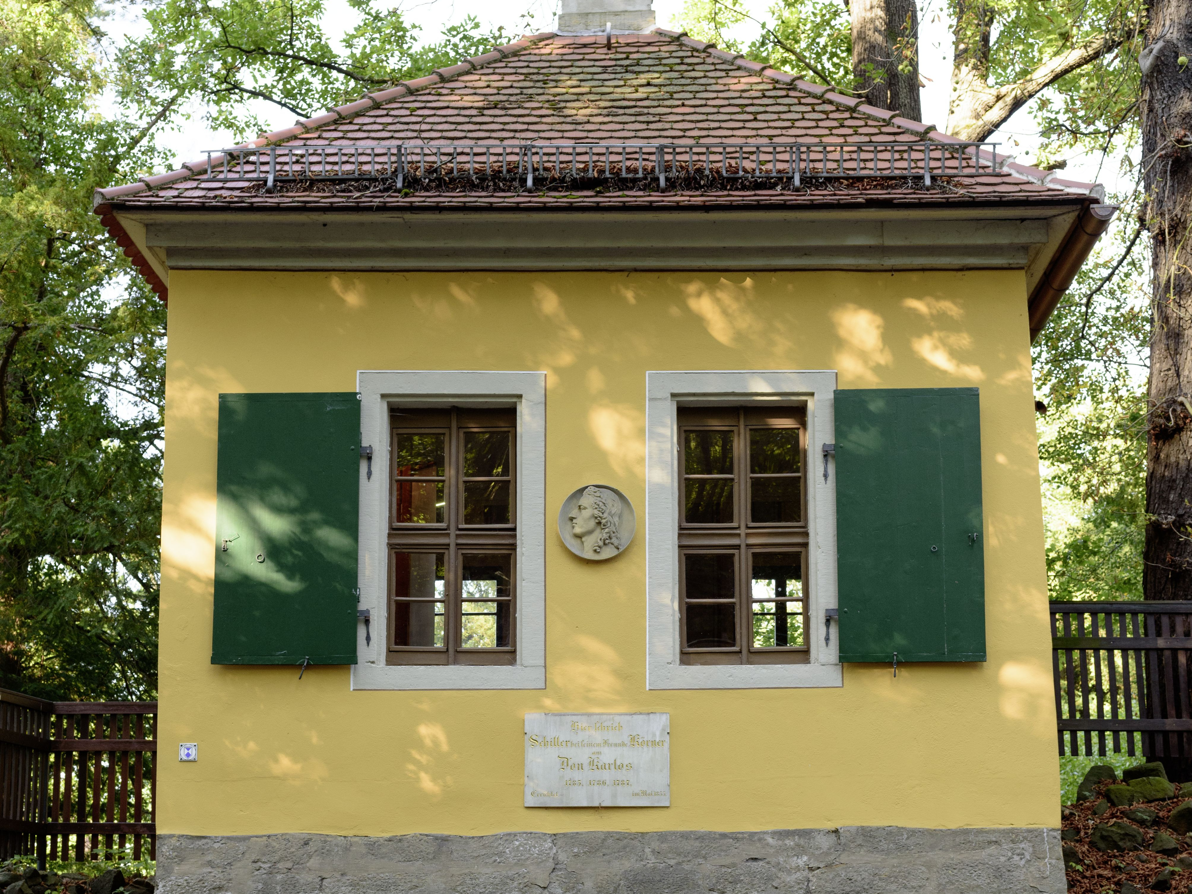 KI generiert: Das Bild zeigt ein kleines, gelbes Gebäude mit grünen Fensterläden, zwei Fenstern und einer Gedenktafel an der Wand. Es ist von Bäumen umgeben, und der Vordergrund wird von einer Steinmauer dominiert.