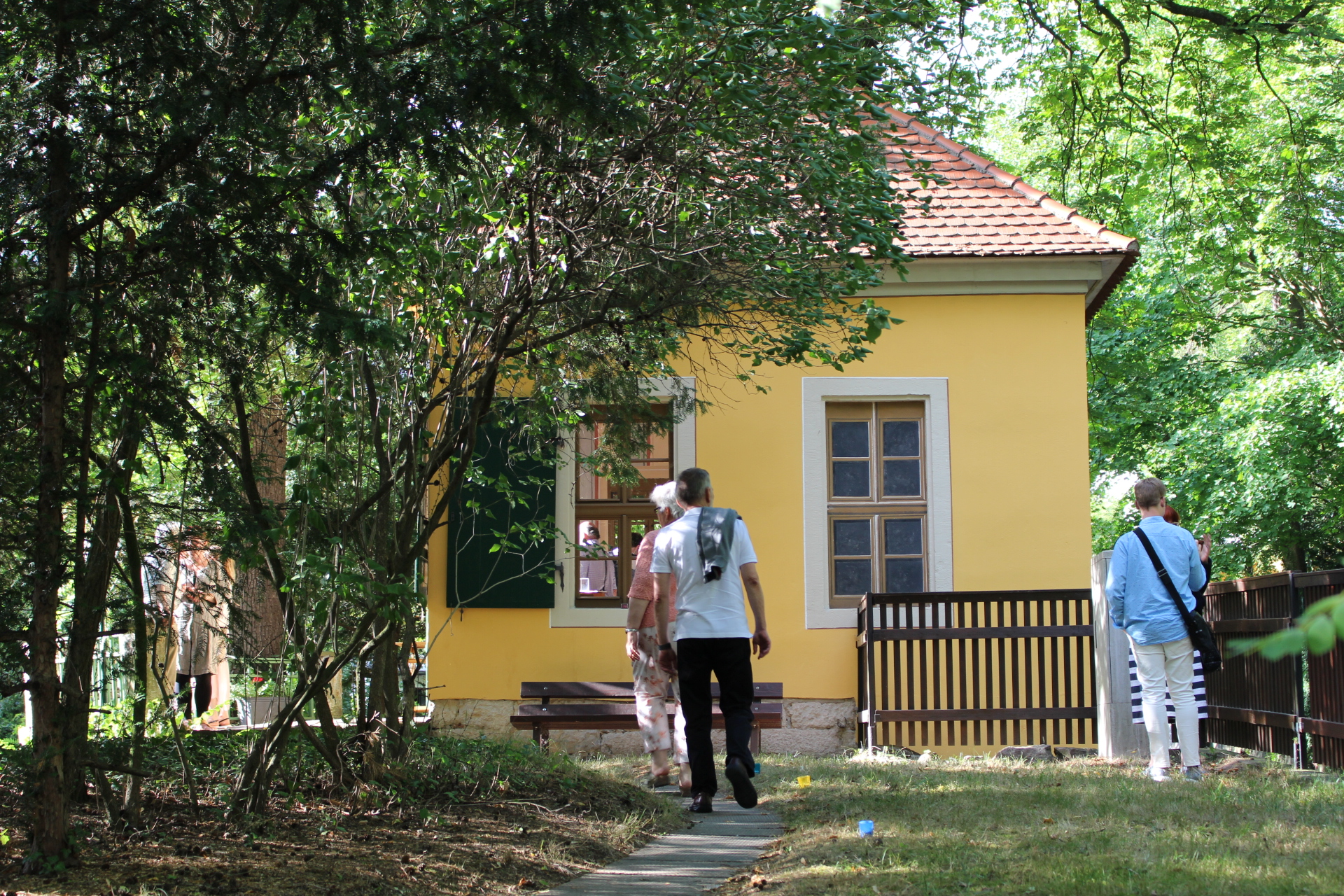 KI generiert: Das Bild zeigt ein kleines, gelbes Haus mit roten Dachziegeln, umgeben von Bäumen und Grünfläche. Im Vordergrund sind mehrere Menschen zu sehen, die das Gelände des Hauses erkunden.