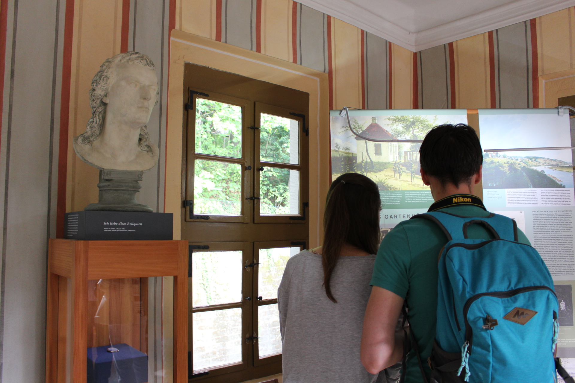 KI generiert: Auf dem Bild sehen wir zwei Personen von hinten, die in einem Ausstellungsraum stehen und sich Informationstafeln ansehen. Links befindet sich eine Büste auf einem Sockel und ein dekoriertes Fenster.